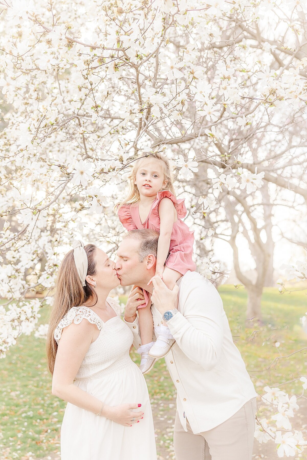 girl sits on dad shoulders white parents kiss during spring maternity photo session in Wellesley Massachusetts with Sara Sniderman Photography