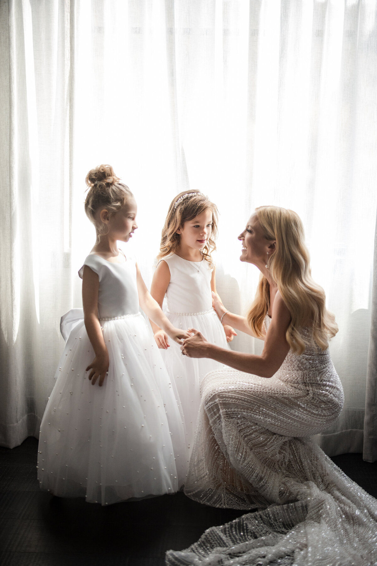 A bride in a white dress kneels beside two young girls in white dresses near a window. This photo was taken by a NYC wedding photographer specializing in iconic venues