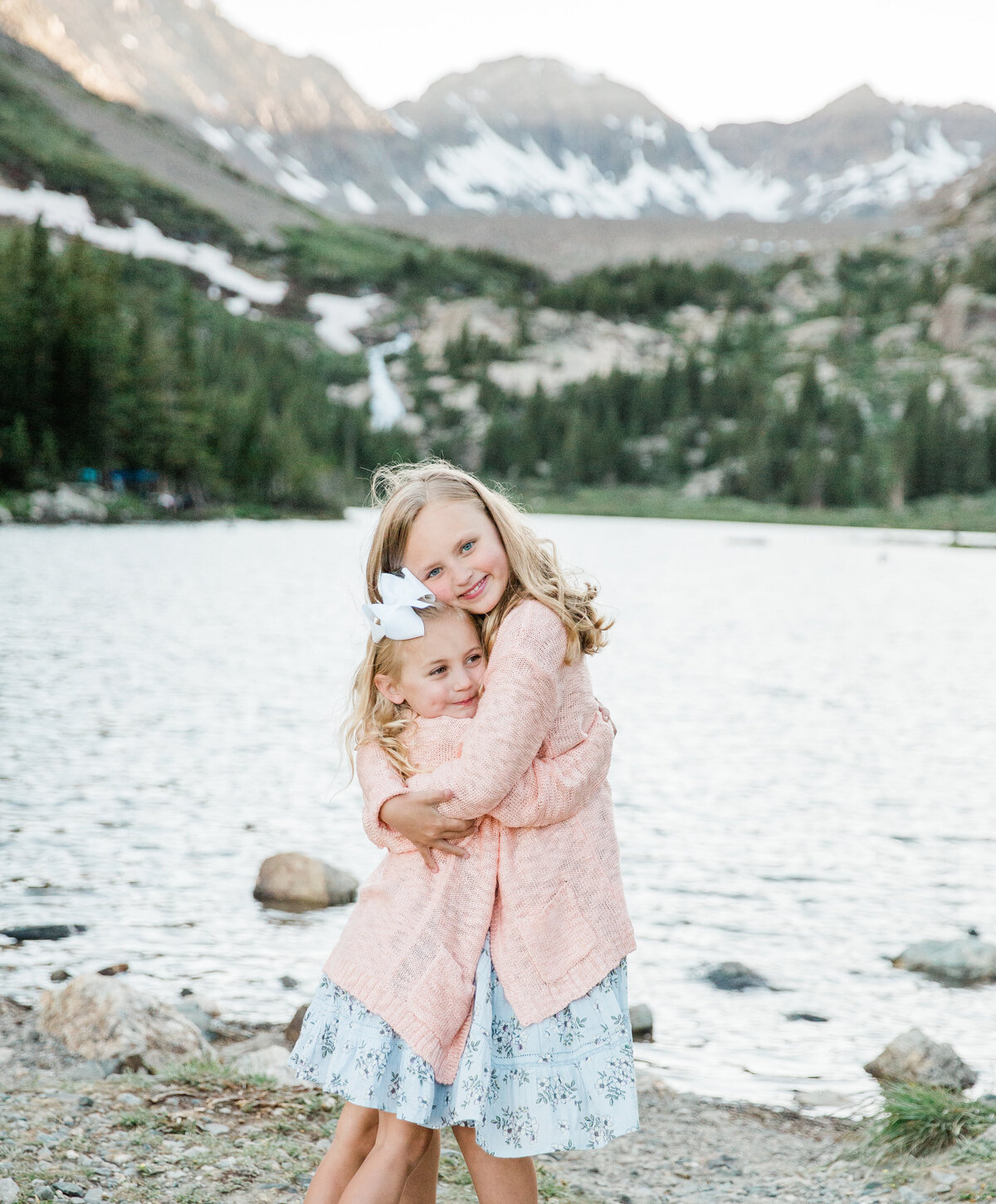 Two young sisters are embracing in a huge on a shore of a lake with large mountains behind them