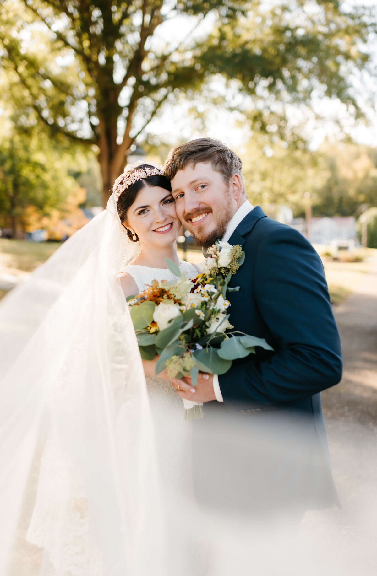 wedding day portrait with bride and groom standing close together with their temples touching and smiling as they both hold onto the bride bouquet and the veil from her head flows in front of them with the sunsetting through the trees behind them captured by Virginia wedding photographer