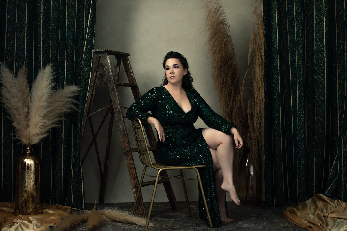 A Caucasian white woman in a green sequin dress sits in a gold chair and looks off camera as as she poses for a professional portrait for Janel Lee Photography studios