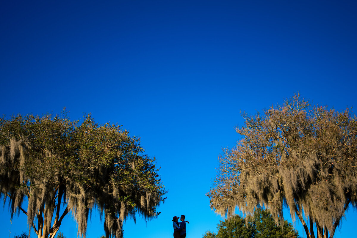 Wide-landscape-image-with-a-big-blue-sky-and-two-mossy-trees-symmetrically-on-each-end-of-the-photograph-with-a-silhouette-of-a-couple-in-the-center-at-Riverfront-Park-in-Columbia-SC