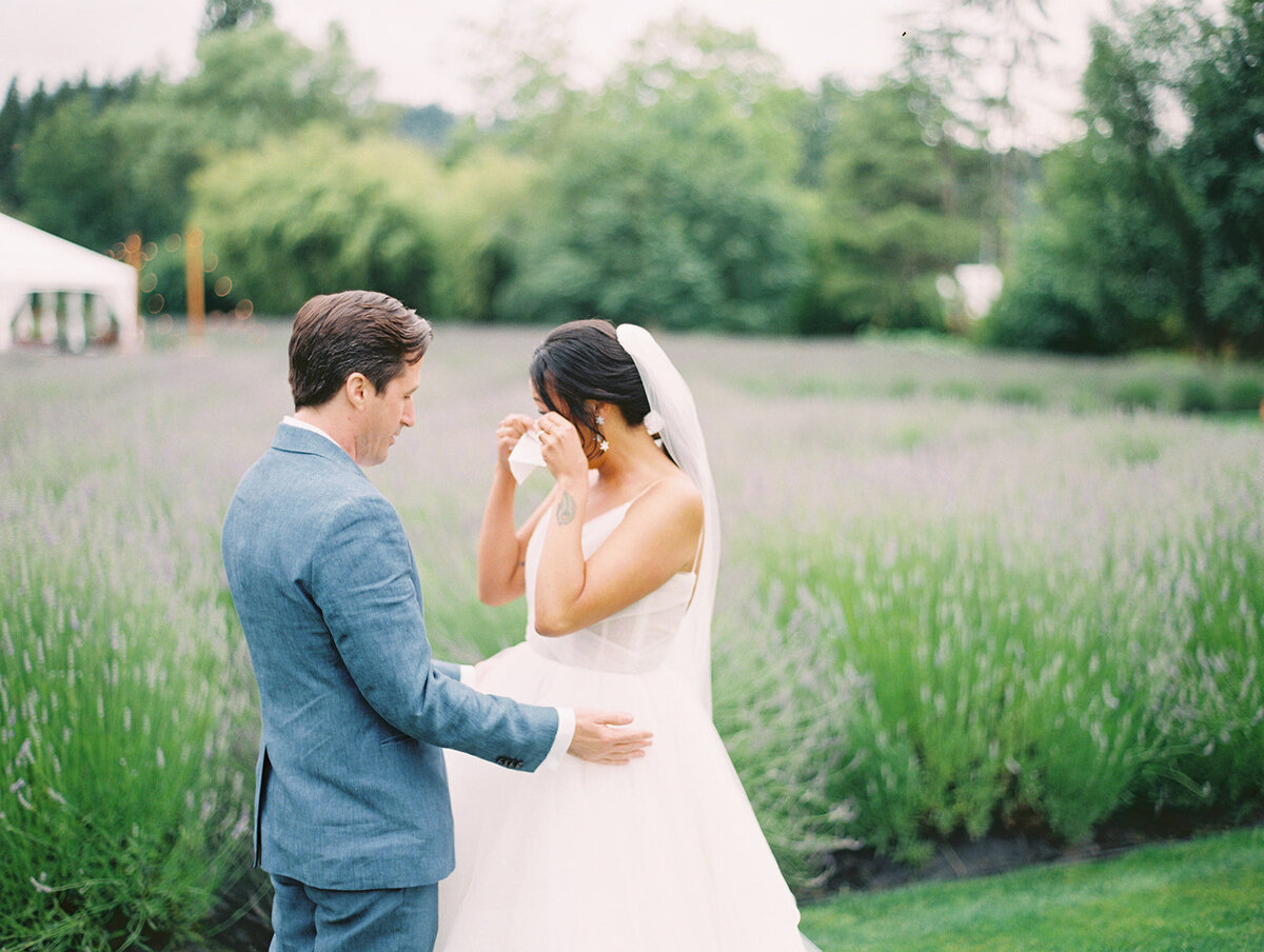 A bride and groom on their wedding day, the bride is holding a tissue to her eyes because she has been tearing up after their first look, and the groom has his arms surrounding him, they are at a lavender farm in san ysidro and the lavender is blooming behind them