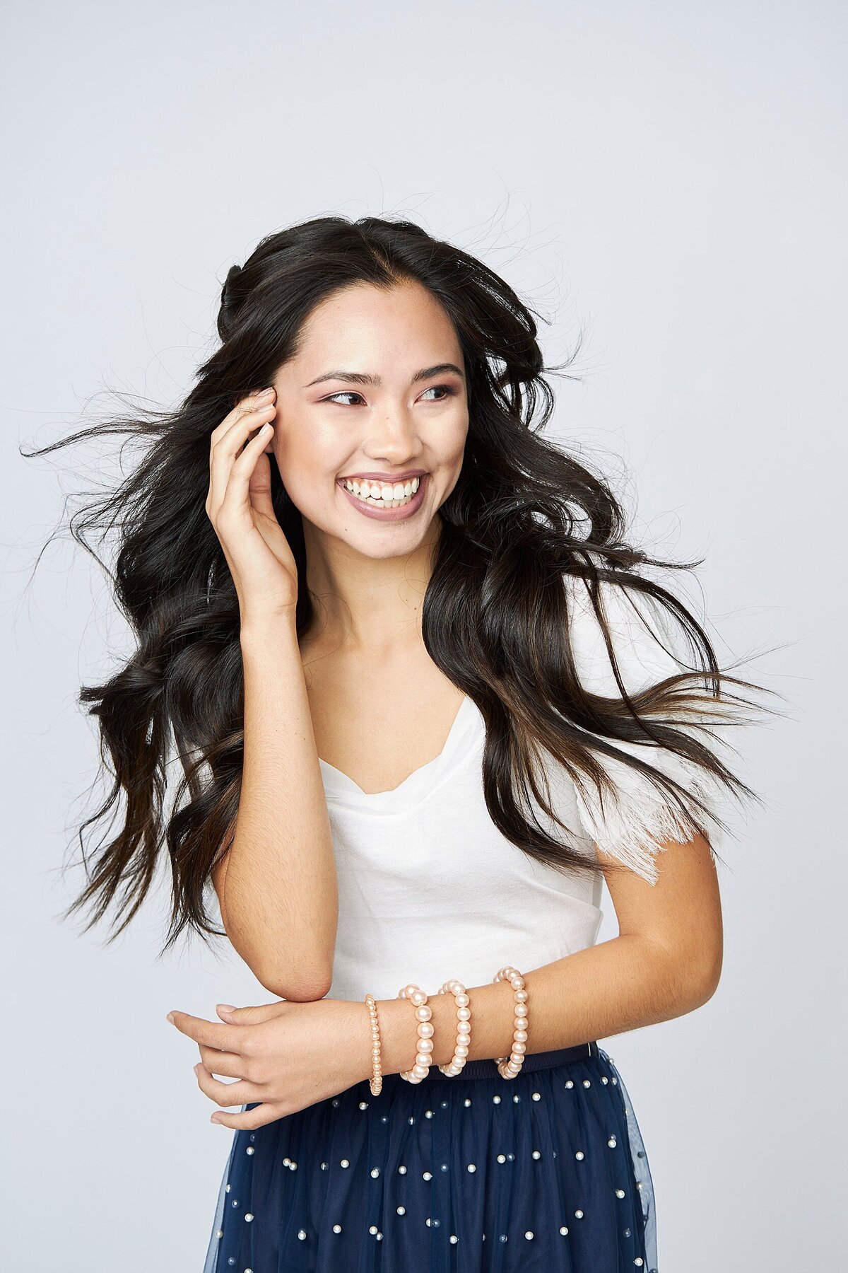 Fun and candid studio photo of a smiling teen girl with long black hair wearing a white top and navy skirt with pearl details.