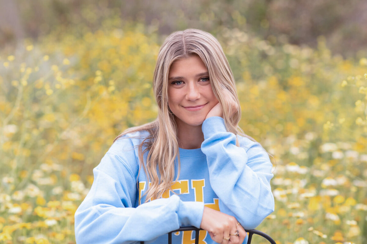 A close-up of a young college student with long, blonde hair wearing a powder blue sweatshirt with yellow lettering on it that says "UCLA," sitting in a black chair and smiling at the camera in a field of yellow flowers at Northwest Open Space in San Juan Capistrano.