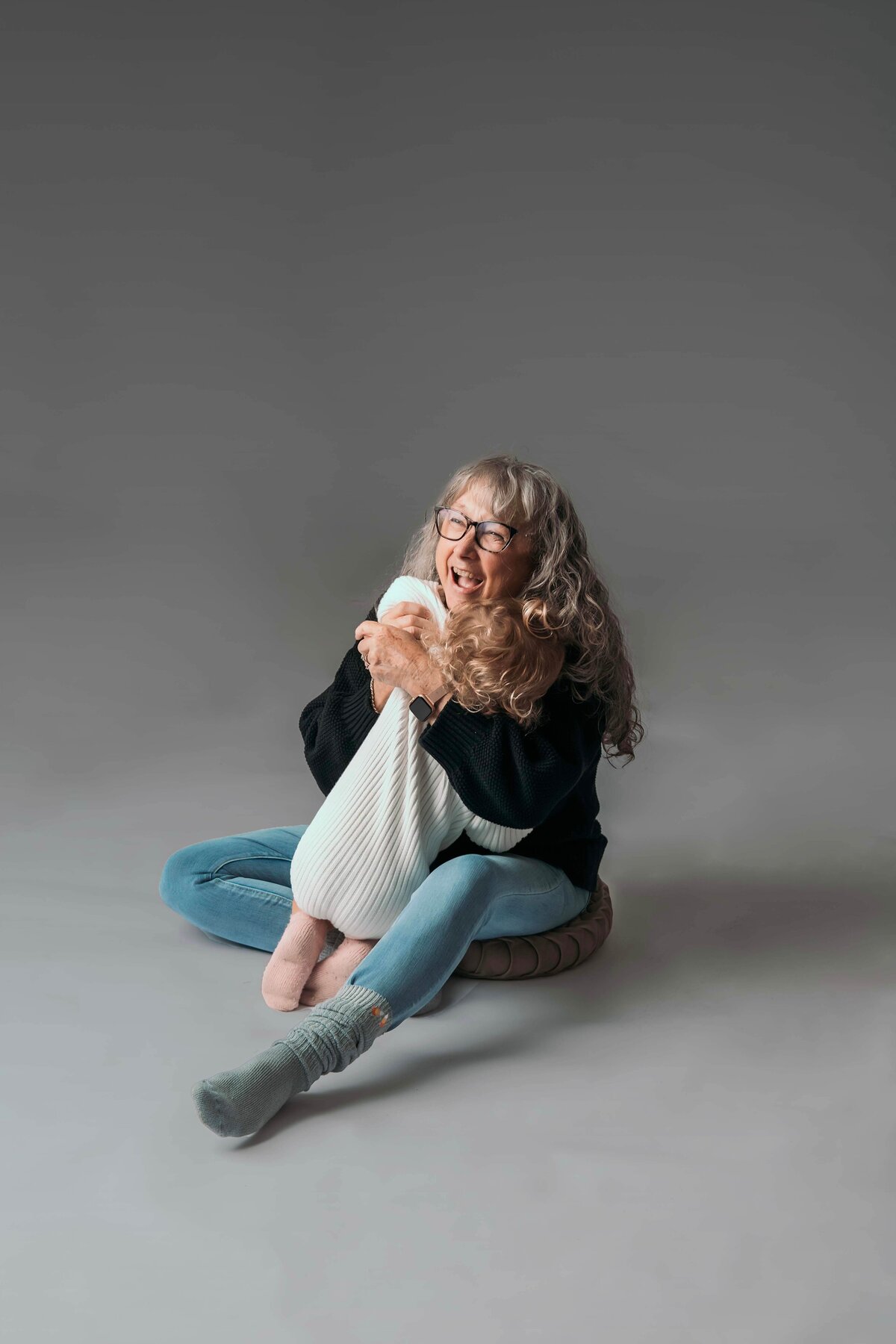 A grandmother dressed in blue jeans, a black sweater, and grey socks sits on the floor of Mobile, AL Motherhood Photographer’s studio, holding her 2-year-old granddaughter in an oversized white sweater onesie. The tender embrace highlights their affectionate bond in this warm and intimate portrait