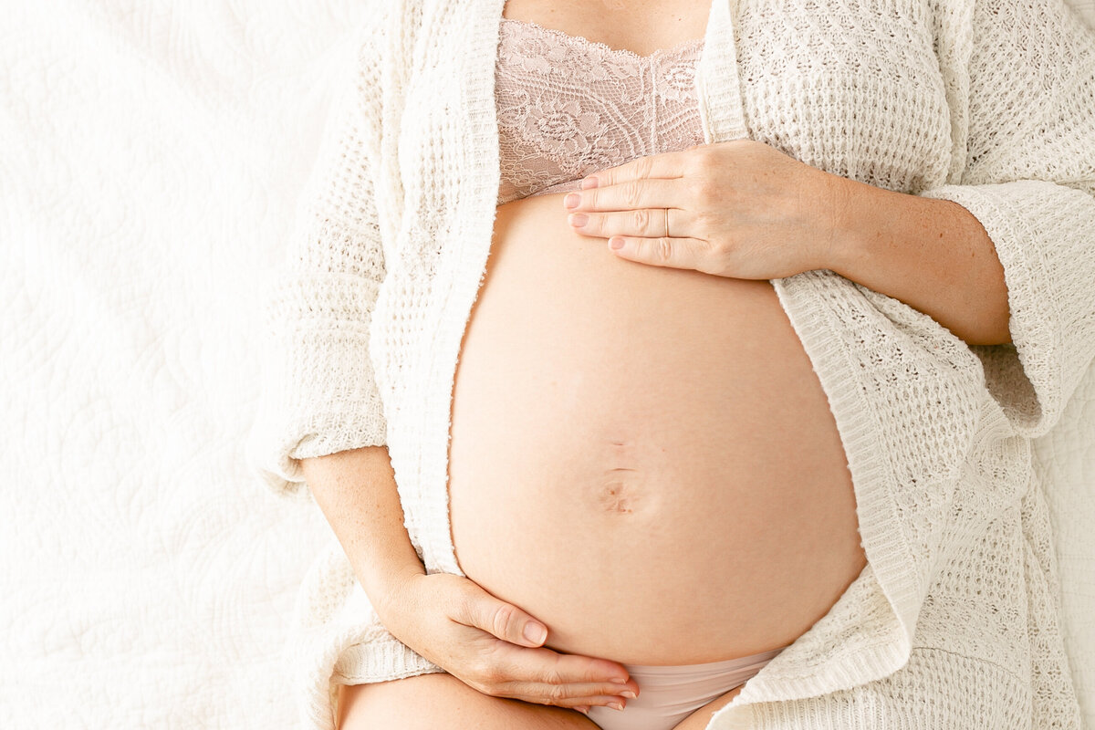 Close-up of bare pregnant belly and woman resting her hands on the top and bottom. She is wearing a blush lace bra and a cream cardigan. Portland Maternity Photographer