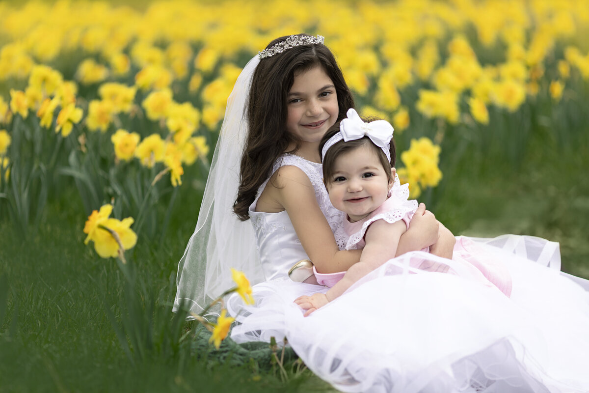 A toddler girl in a pink dress plays in her big sisters lap in a field of daffodils