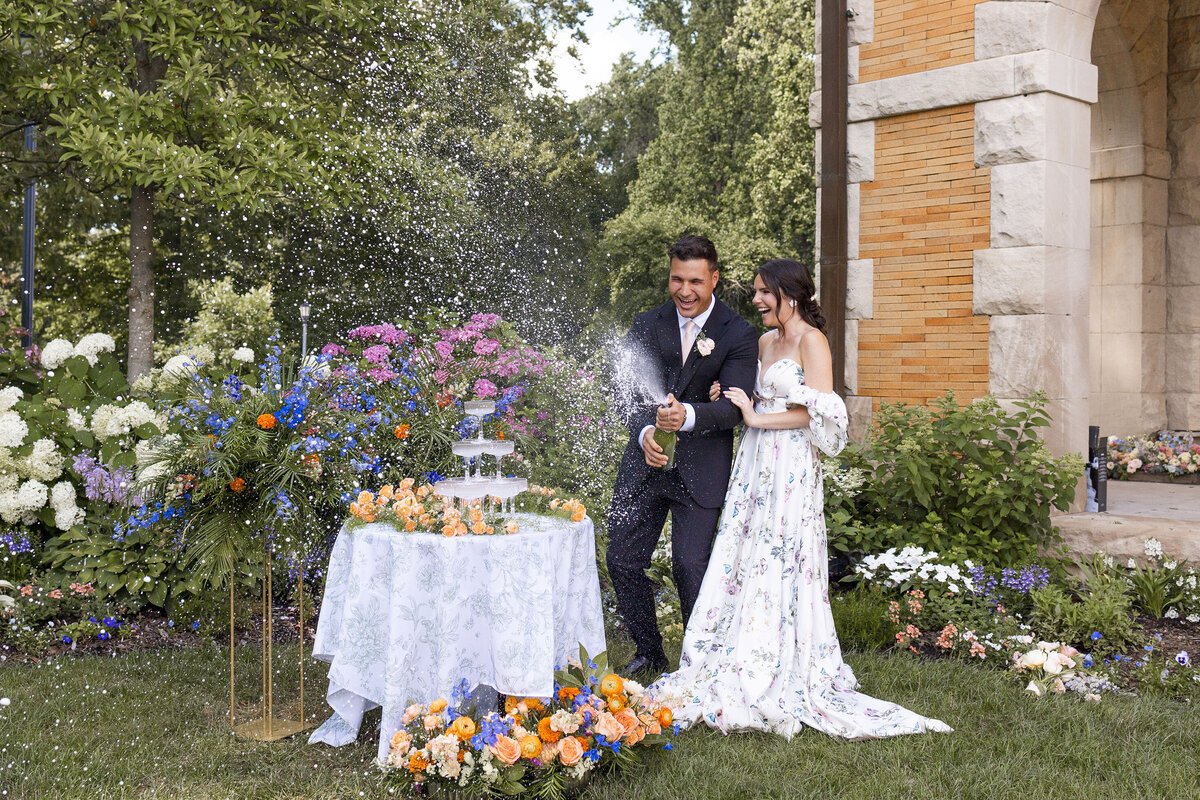 Whimesical picture of a Groom doing a champagne spray with the champagne tower in beautiful garden setting surrounded by florals and his joyful bride.