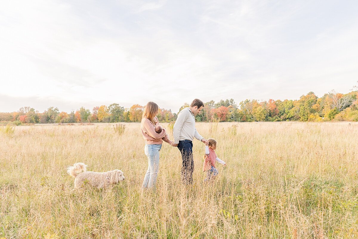 family walks through field  during outdoor newborn photo session with Sara Sniderman Photography in Natick Massachusetts