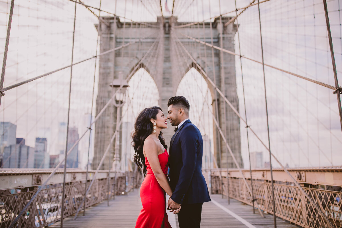 An engaged couple in red formal outfit hold hands while on the brooklyn bridge in nyc