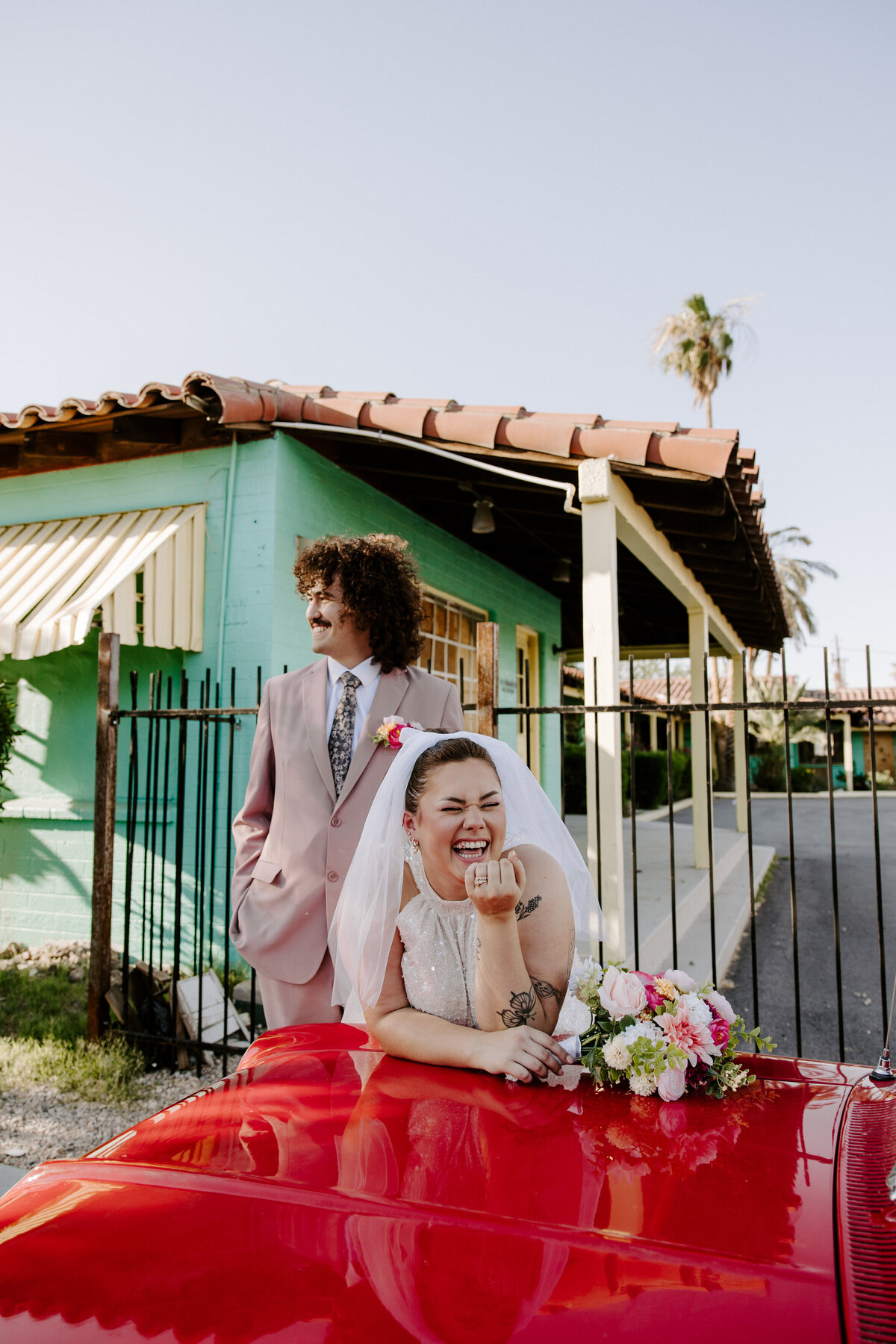 A bride smiles while holding flowers while their partner looks to the side, also laughing.