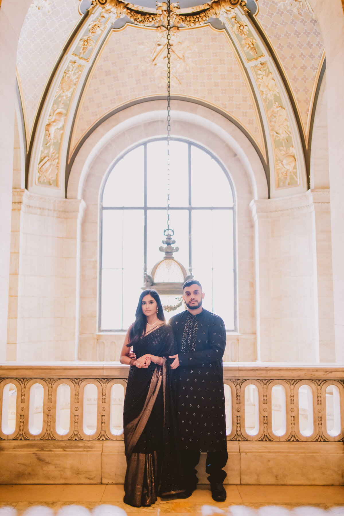 An engaged couple standing in front of a window in indian outfits at the new york public library in indian outfits