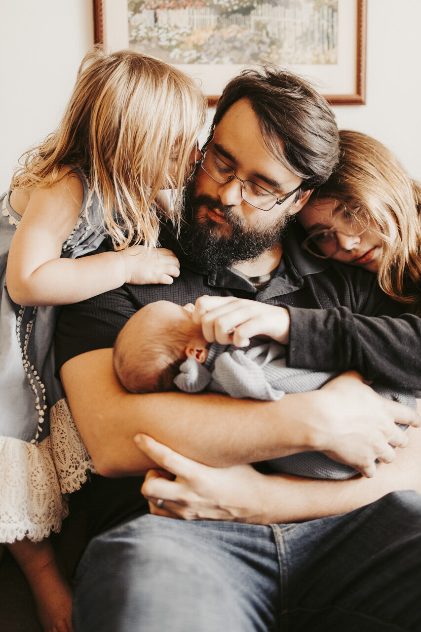 Sweet picture of Dad holding newborn, with big sisters on either side of him, one kissing his cheek, the other laying her head on his shoulder: lifestyle newborn photography in Tifton