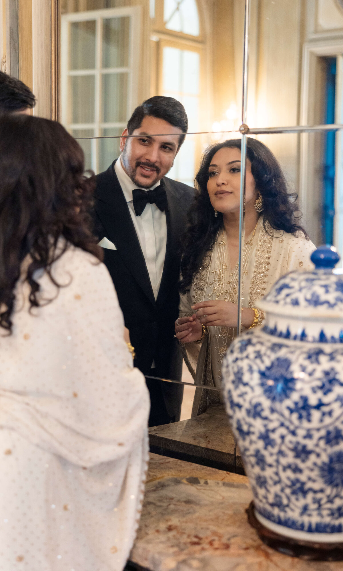 A man in a black tuxedo and a woman in a white embellished outfit admire themselves in a mirror. They stand next to a blue and white decorative vase. The setting seems elegant, with soft lighting and a refined interior.