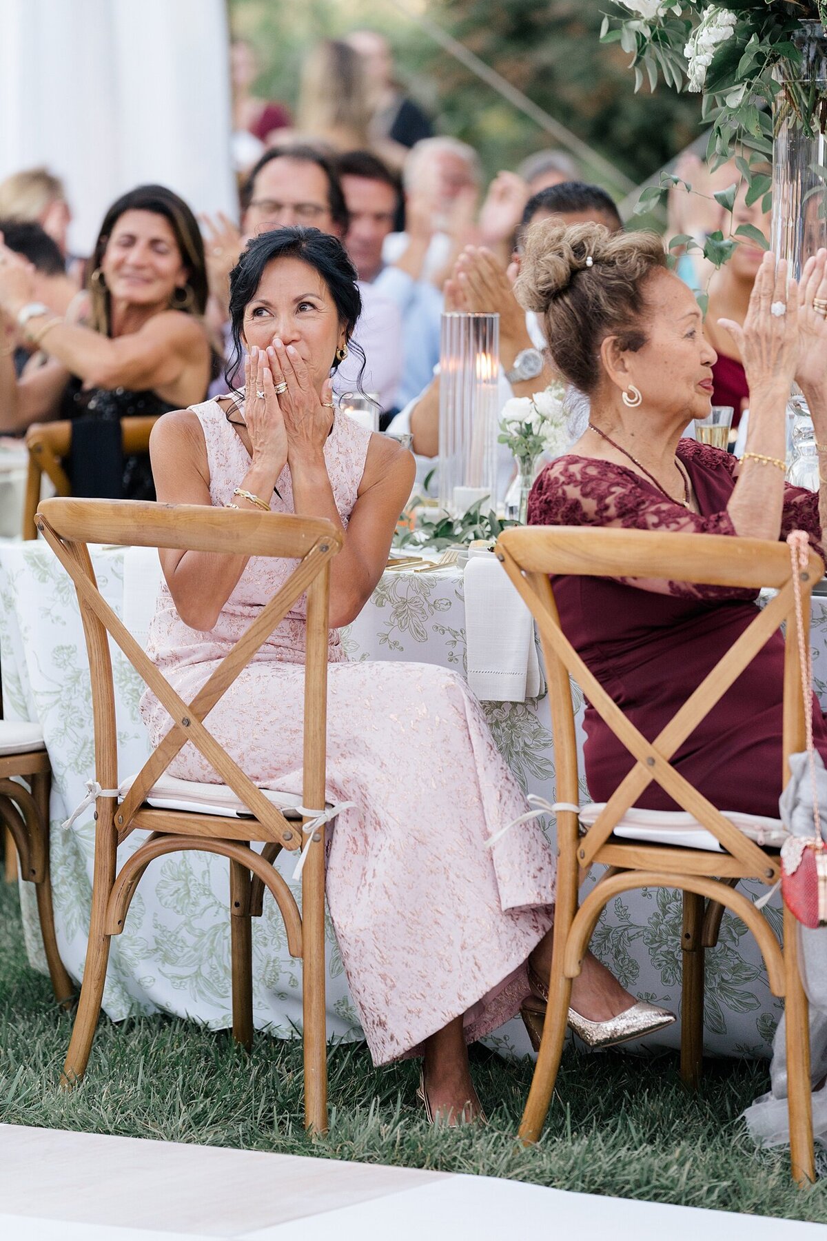 wedding guests sitting on wooden rental chairs in the grass of the backyard wedding under a white tent
