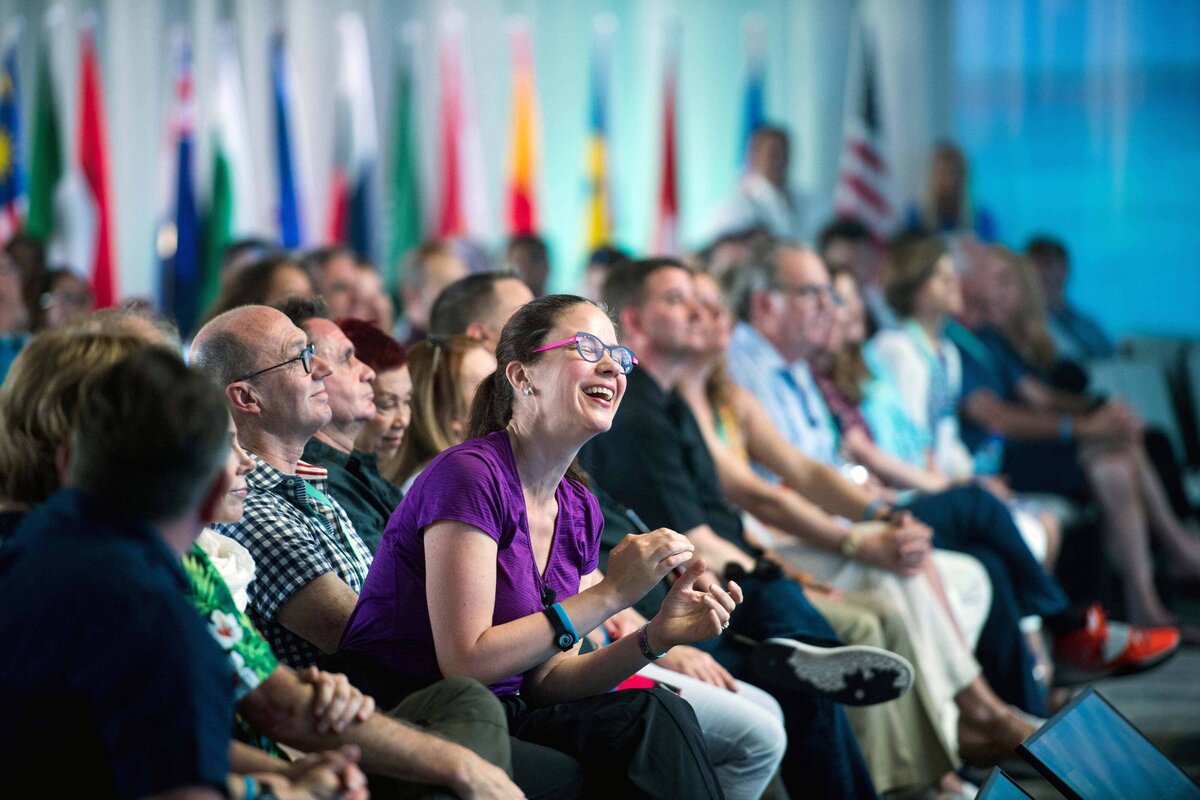 Women leans forward laughing at international conference while seated in audience with various flags in background