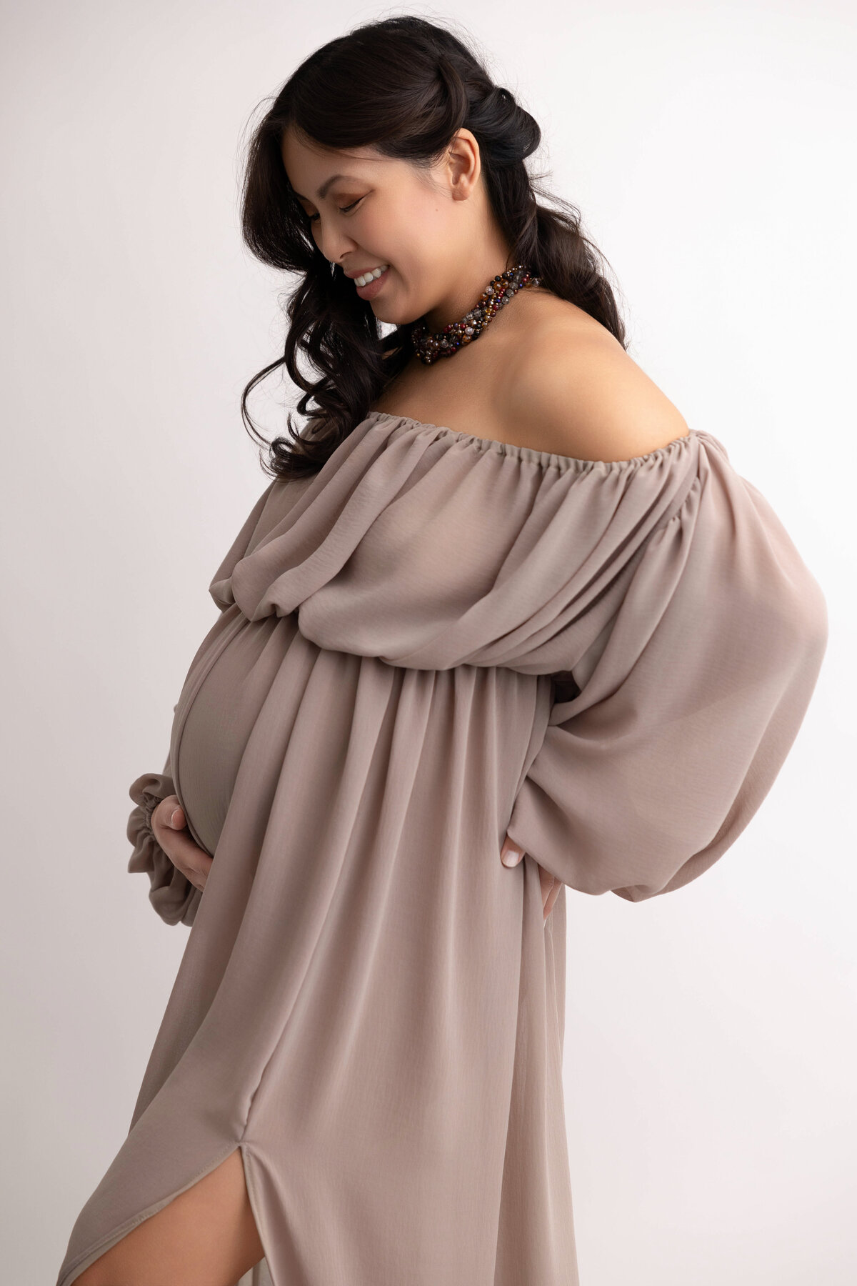 Pregnant mother wearing a light brown flowing dress and smiling down at pregnant belly while holding with one hand under her belly.