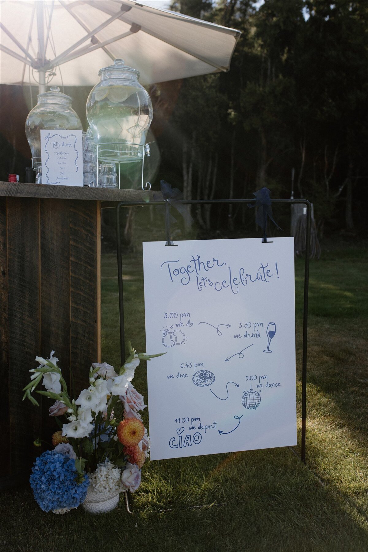 Hand-drawn wedding timeline sign at a relaxed alfresco celebration in Wanaka, displayed beside citrus water dispensers and wild floral arrangements.