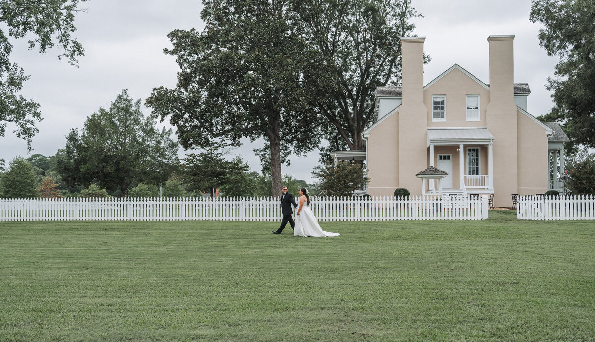 A newlywed couple walks hand-in-hand across a vast green lawn with a white picket fence, in front of the historic Windsor Castle Manor in Smithfield, Virginia, captured by Quinn Photography LLC.