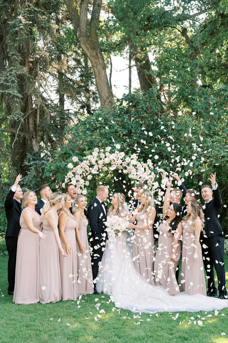 A newlywed couple stands under an arch of flowers, surrounded by their wedding party in taupe dresses and black suits, as flower petals are thrown in celebration, all made possible with the help of expert wedding planning services.