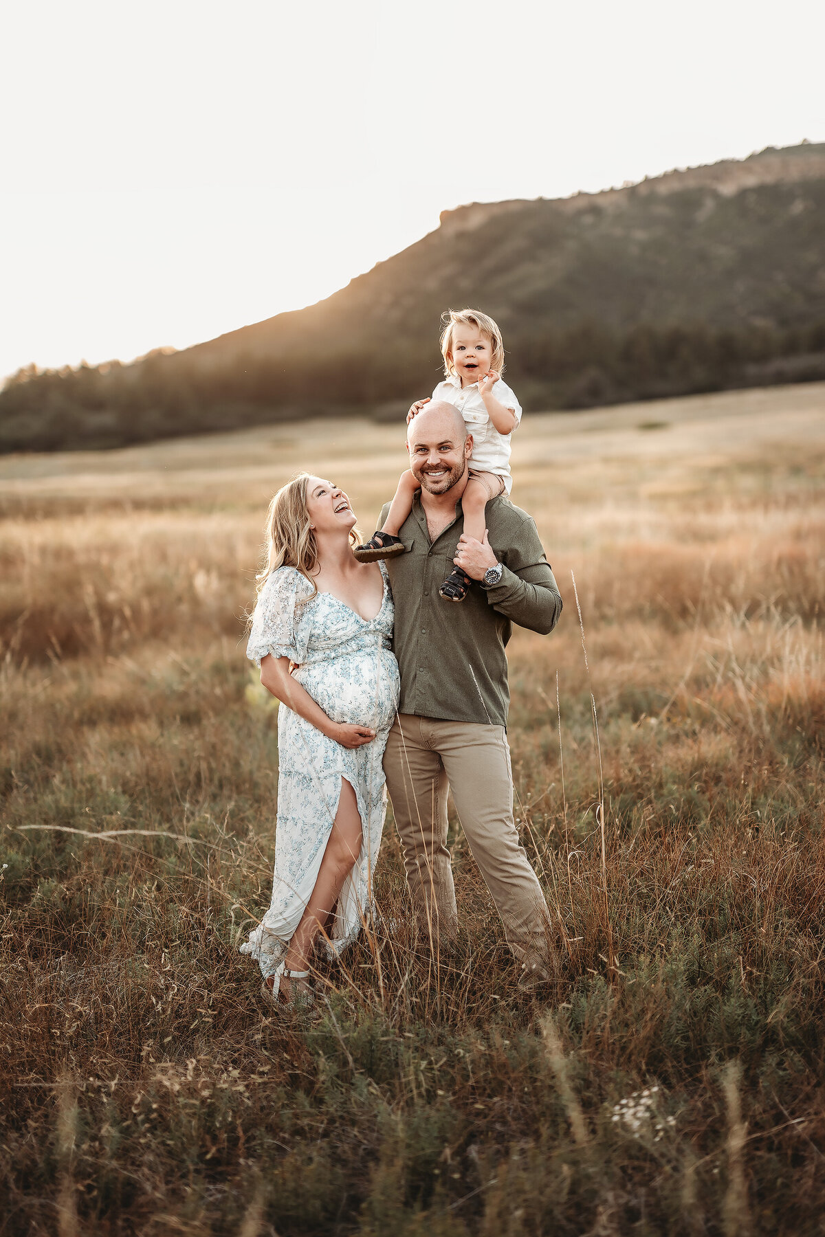 family in a field with boy on dad's shoulders and pregnant mother looking at boy