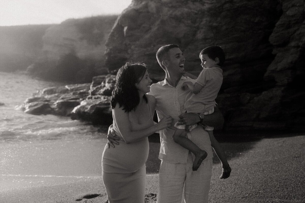 Papa souriant, portant son fils dans ses bras et accompagné de la maman sur une plage rocheuse. Contre-jour doux. Séance grossesse en famille par Kamescop.