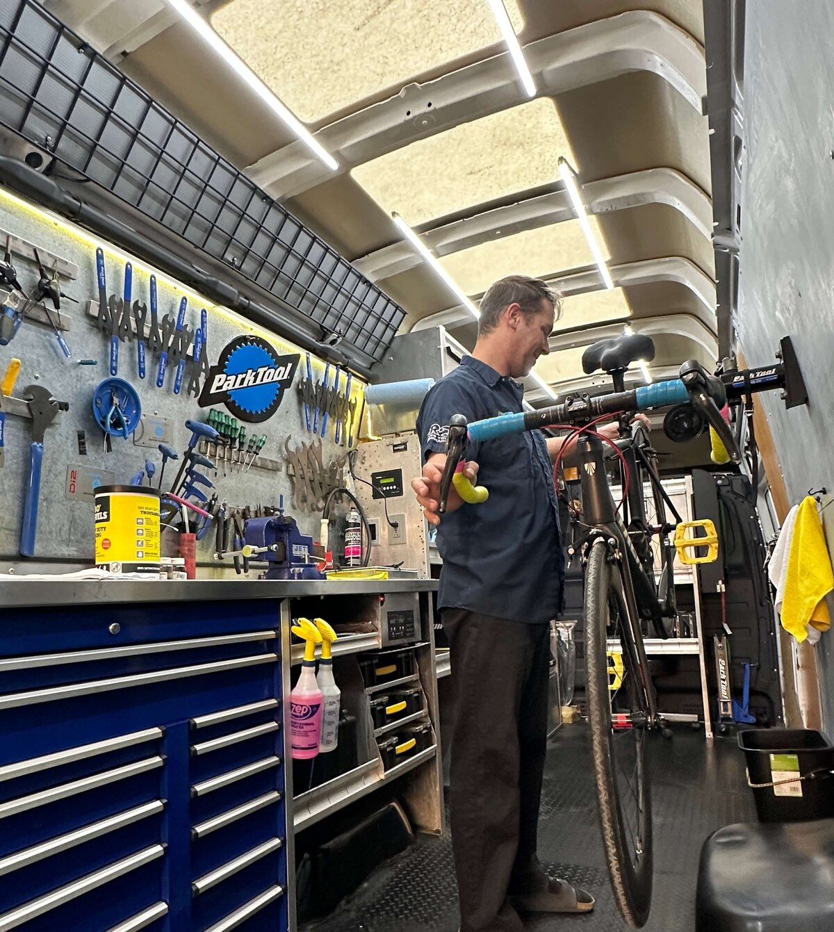 A person working on a bicycle in a well-equipped workshop inside a vehicle. Tools and supplies are organized on the walls and workbench. The person is adjusting the bike on a stand, surrounded by various maintenance equipment and accessories