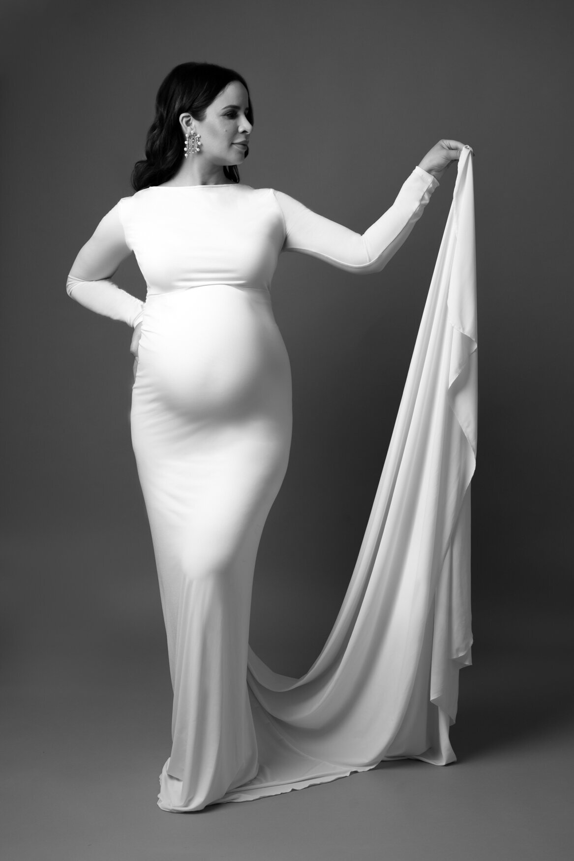 Black and white maternity portrait of a woman in a long-sleeved, form-fitting white gown, elegantly holding the flowing fabric with one hand.