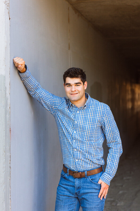 Boy in blue shirt leans against wall while posing in a tunnel for his senior photos.