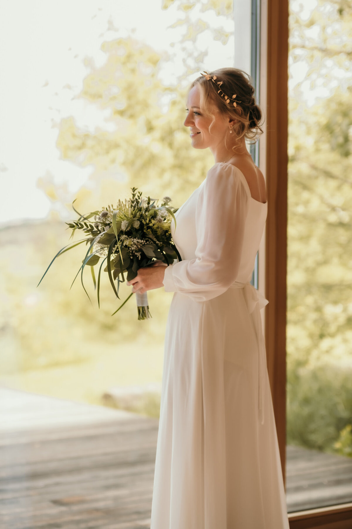 Fertig gestylt blickt die Braut mit ihrem Brautstrauß in den Händen aus einem bodentiefen Fenster.