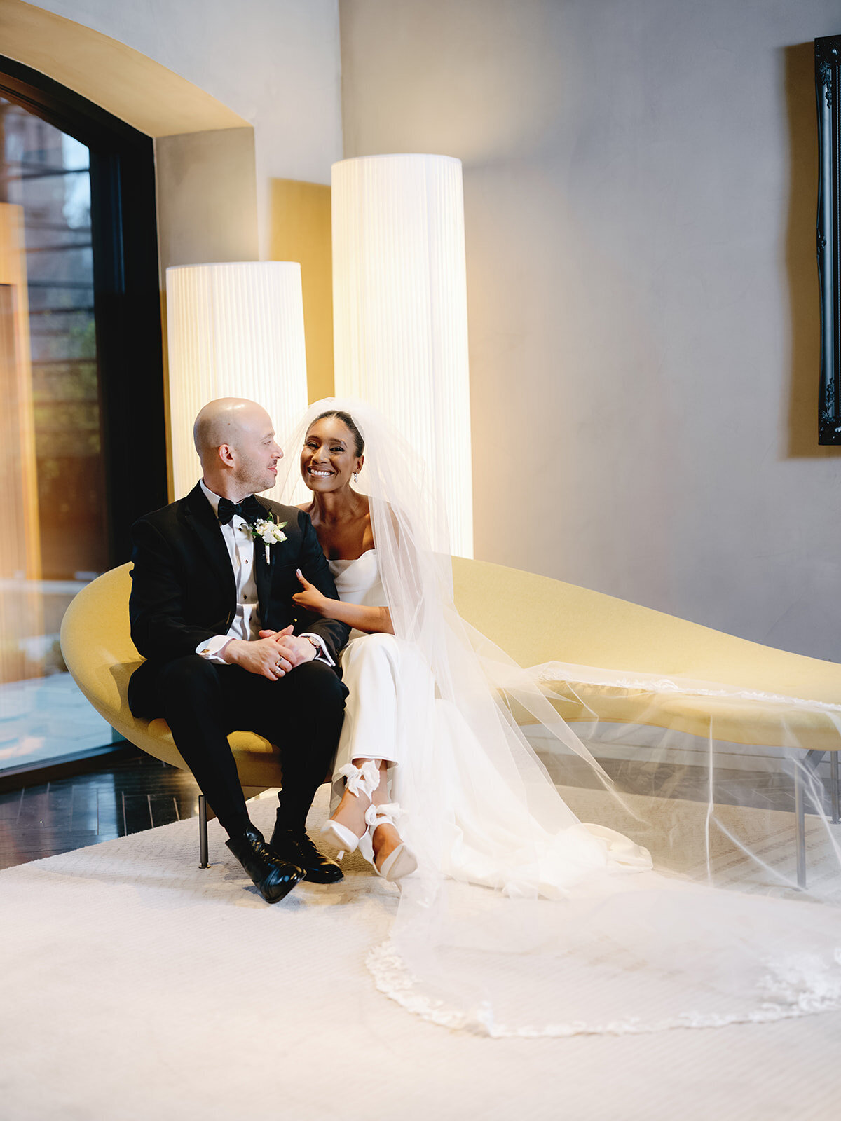 Newly married couple smile as they sit on a yellow couch at their Hotel AKA wedding. 