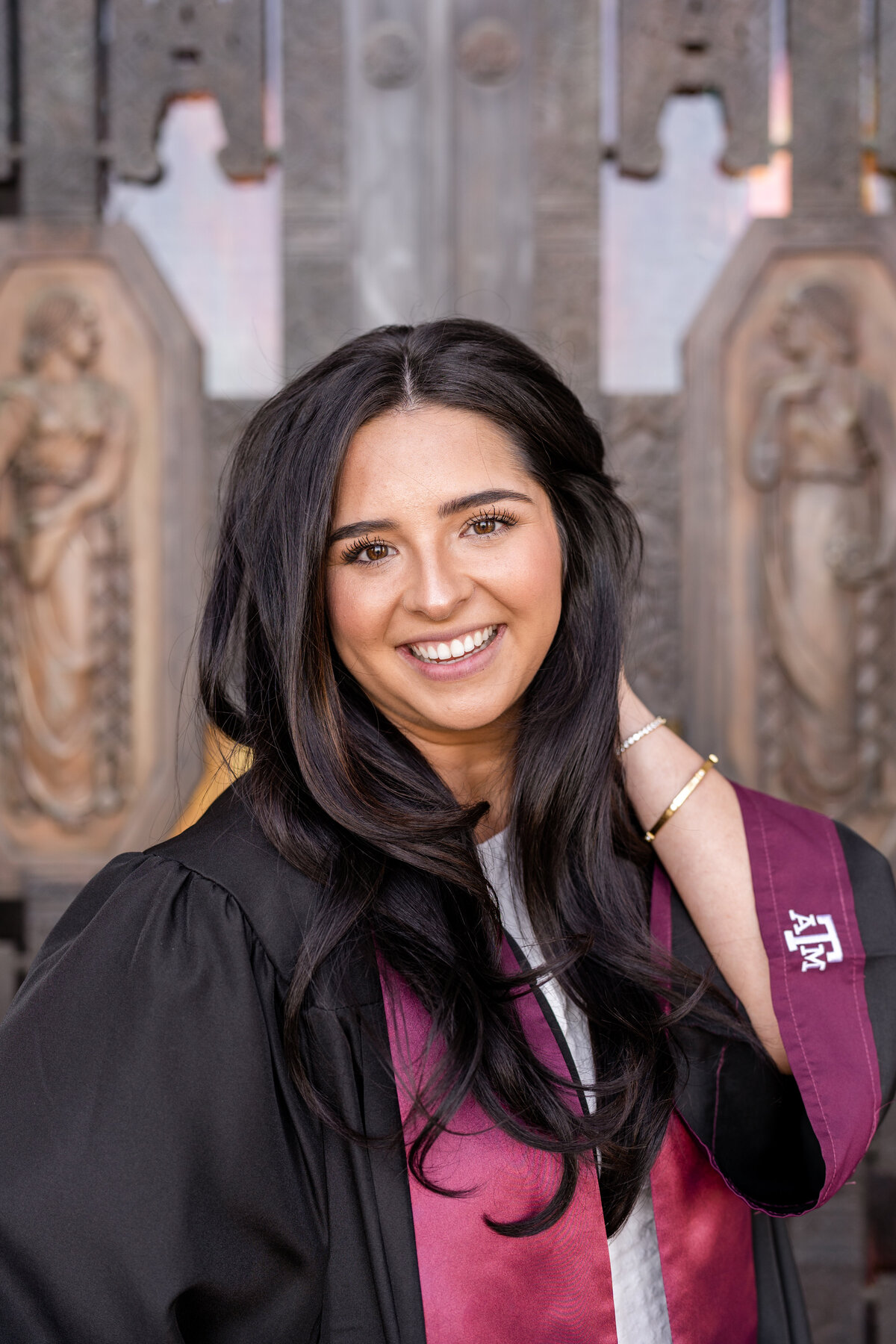Texas A&M senior girl with hand in hair while wearing gown and maroon stole and smiling while standing in front of Administration Building front doors