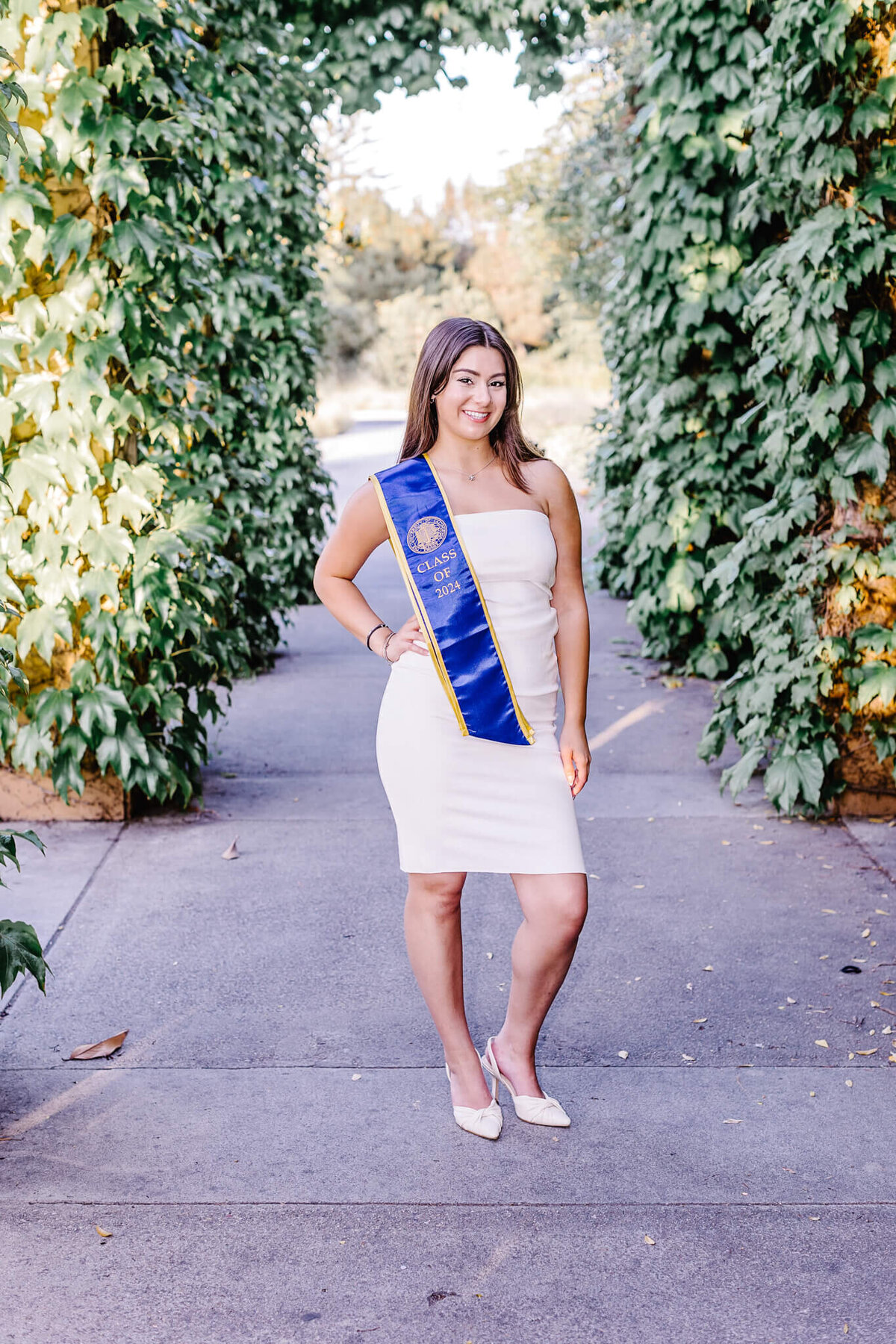 A woman stands on a tree-lined path wearing a white dress and a blue graduation sash. She smiles at the camera, surrounded by lush greenery.