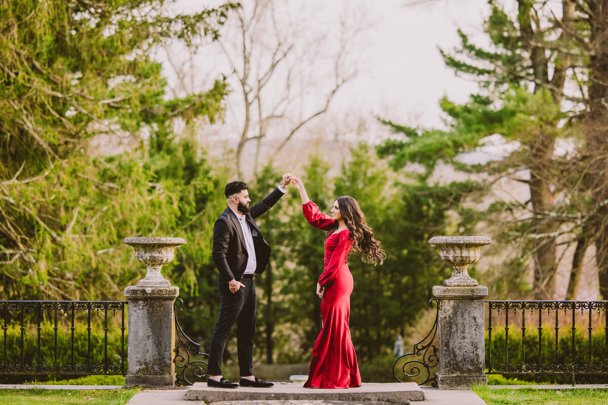 an engaged couple wearing formal red outfits twirling at the nj botanical garden at ringwood state park