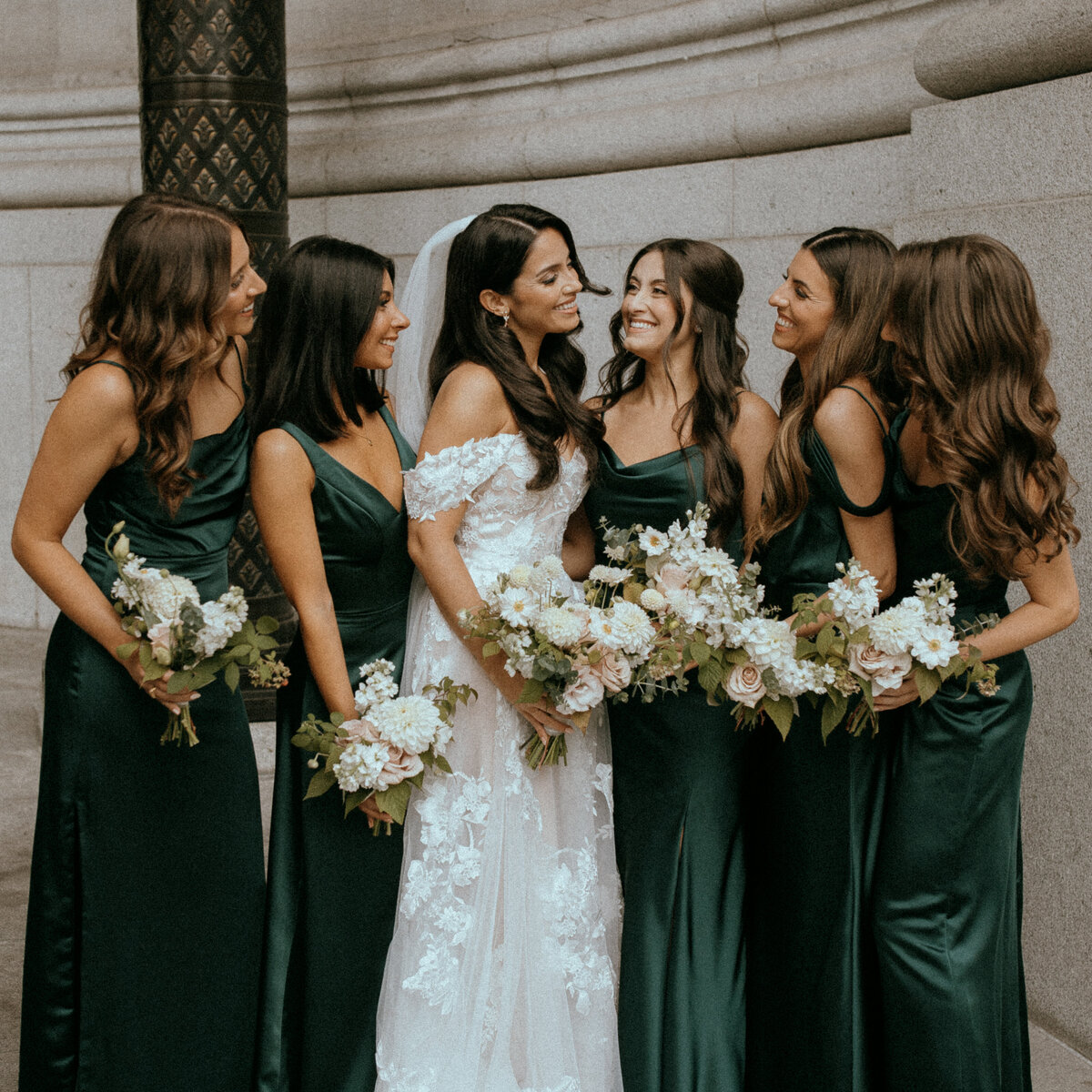 A bride and bridesmaids wearing dark green dresses pose in Old Montreal, holding bouquets.