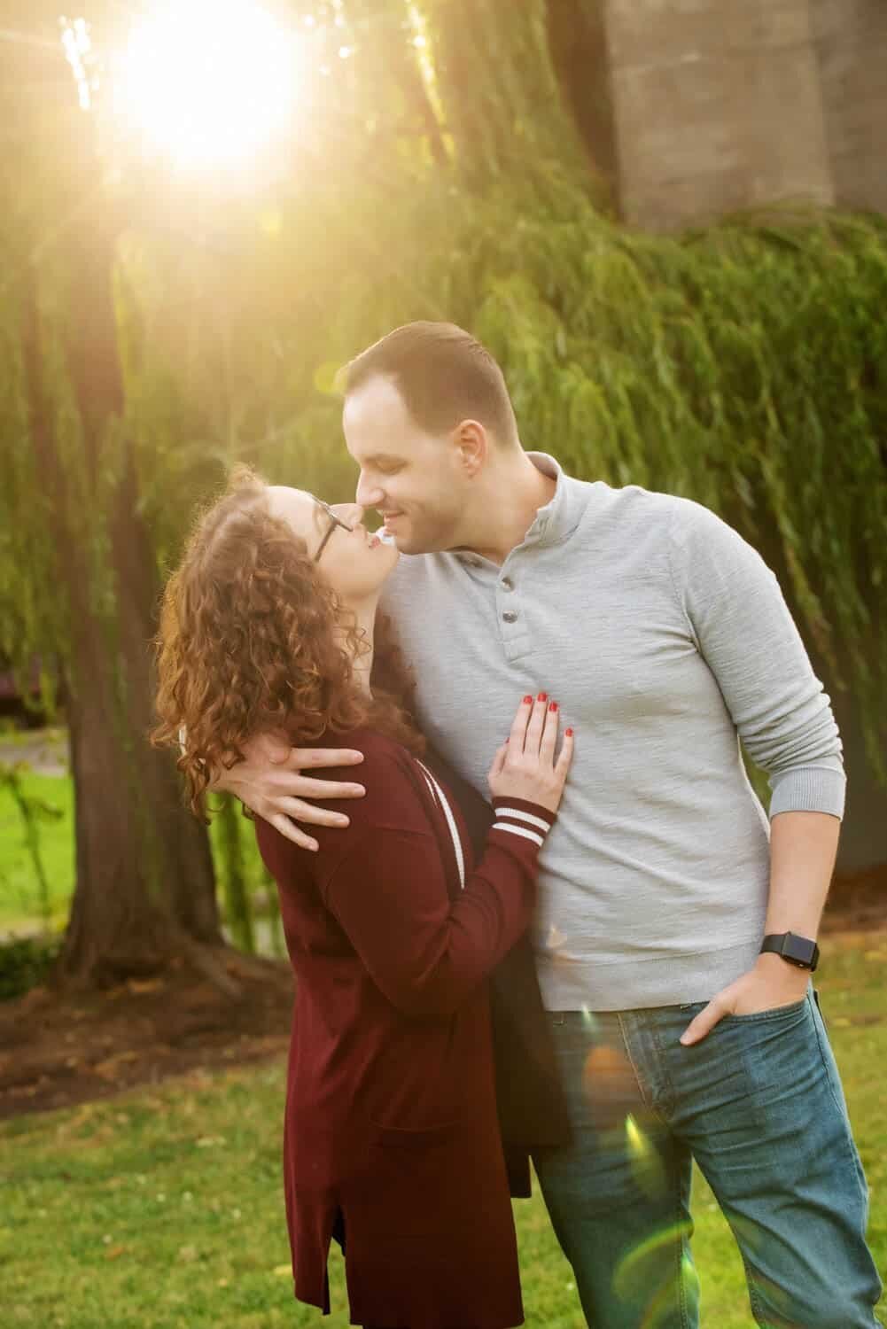 a man and woman touch noses standing in a ray of sunlight in front of a weeping willow