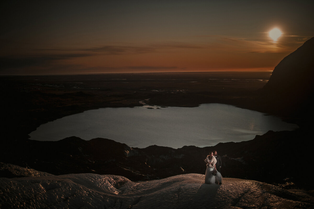 A couple standin gon a rock formation overlooking a large lake a sunset.