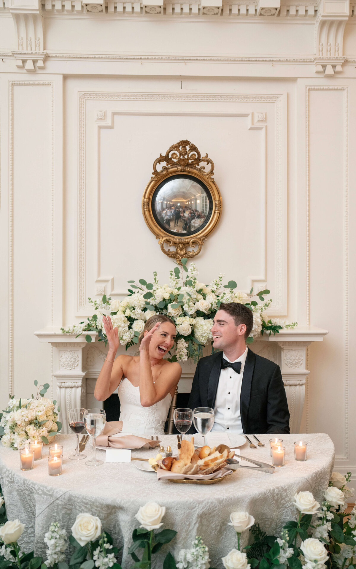 A bride and groom sit at a decorated table in an elegant room with white floral arrangements and candles. The bride claps joyfully, and both are smiling. A round mirror and ornate molding adorn the wall behind them.