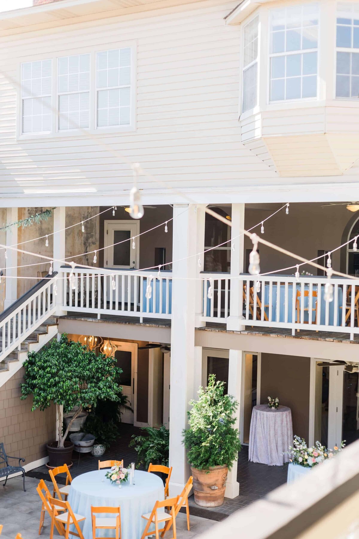 A courtyard with round tables covered in light blue tablecloths, wooden chairs, potted plants, and string lights, surrounded by a second-story balcony with white railings and staircases.