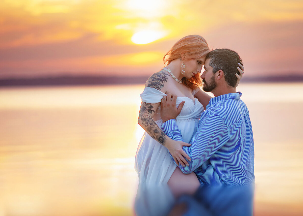 A pregnant couple snuggles in the water on a beach at sunset