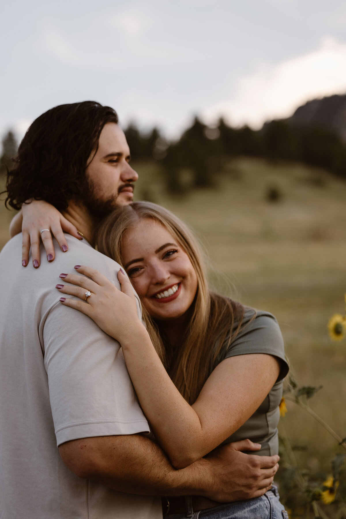 A woman smiles brightly while hugging her partner from the side, showing off her engagement ring with a mountain field in the background.