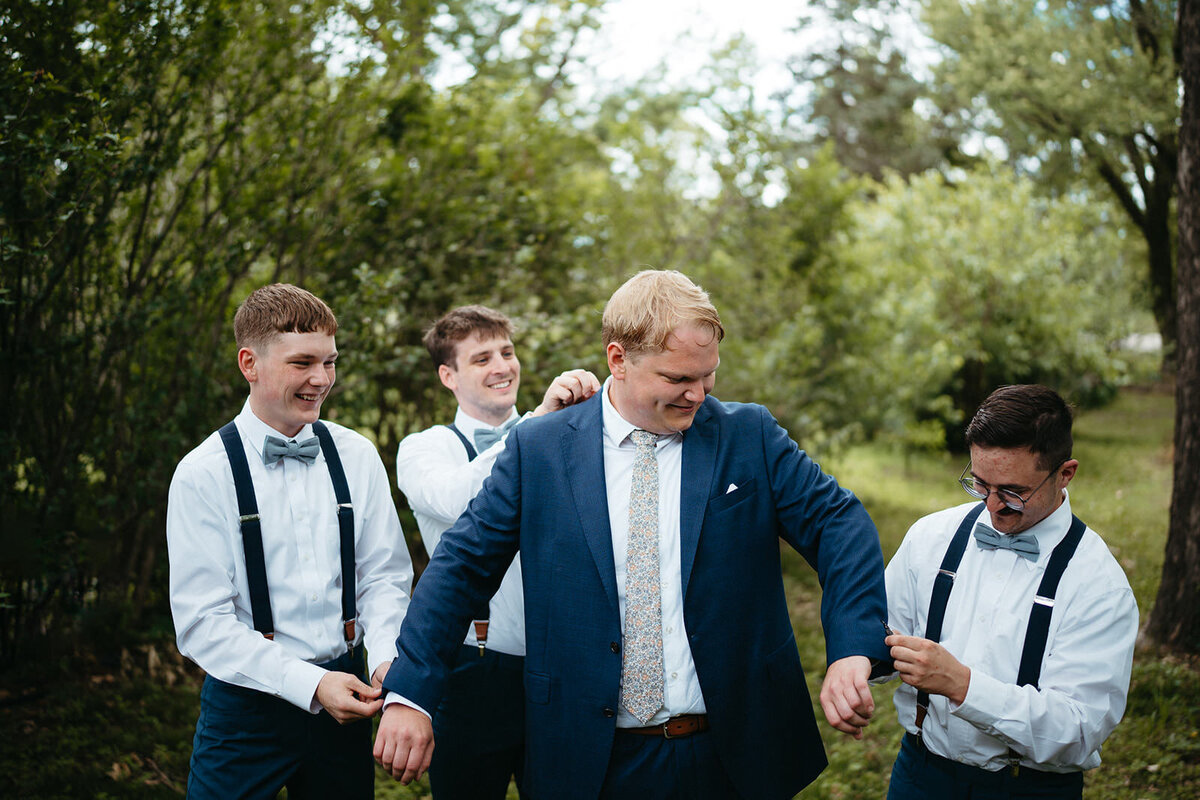 Three people helping a newlywed put on a suit jacket 