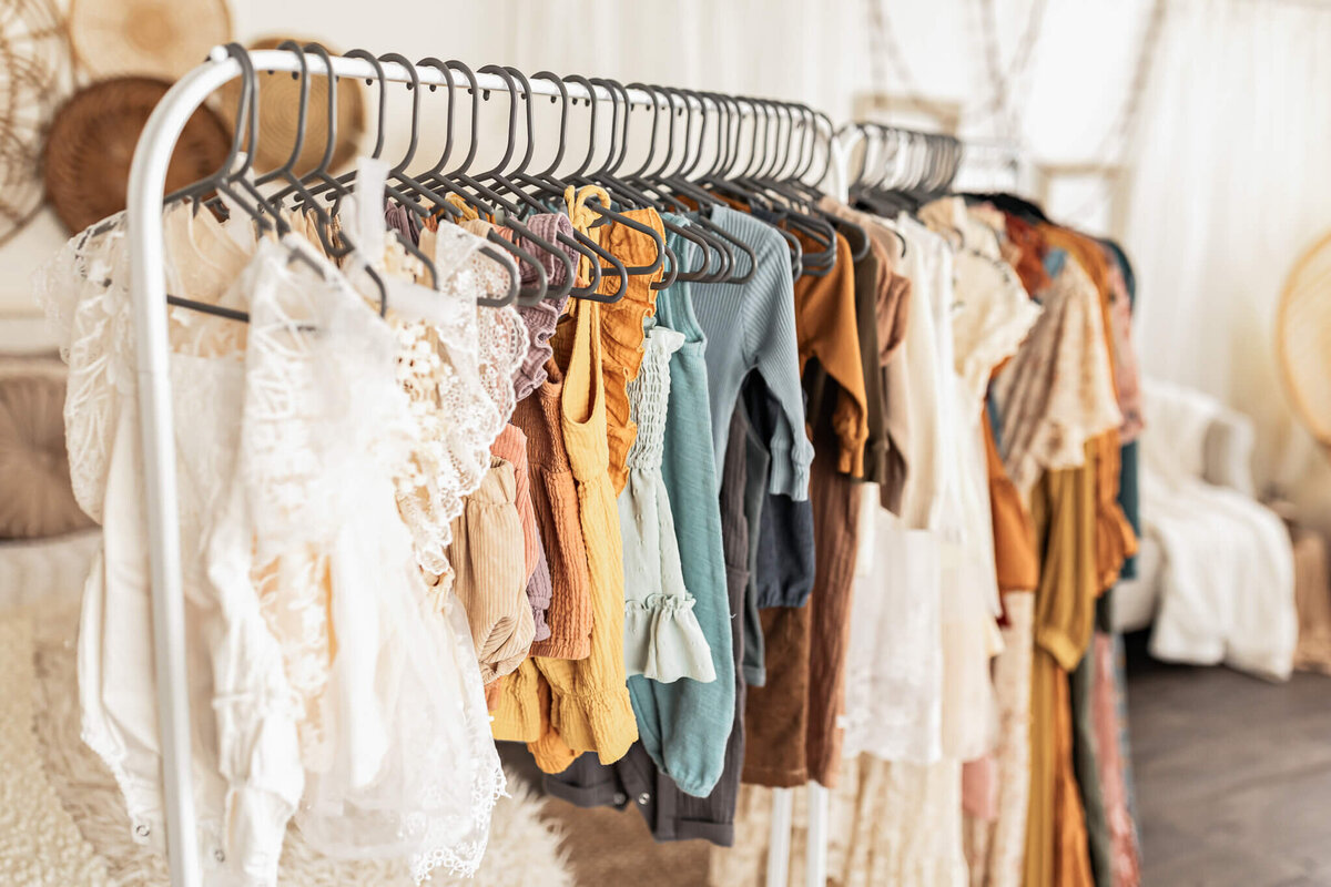 A clothing rack filled with colorful Victorian-style garments, including blouses and dresses with ruffles and lace. The background features soft, neutral-toned decor, creating a cozy, vintage atmosphere.