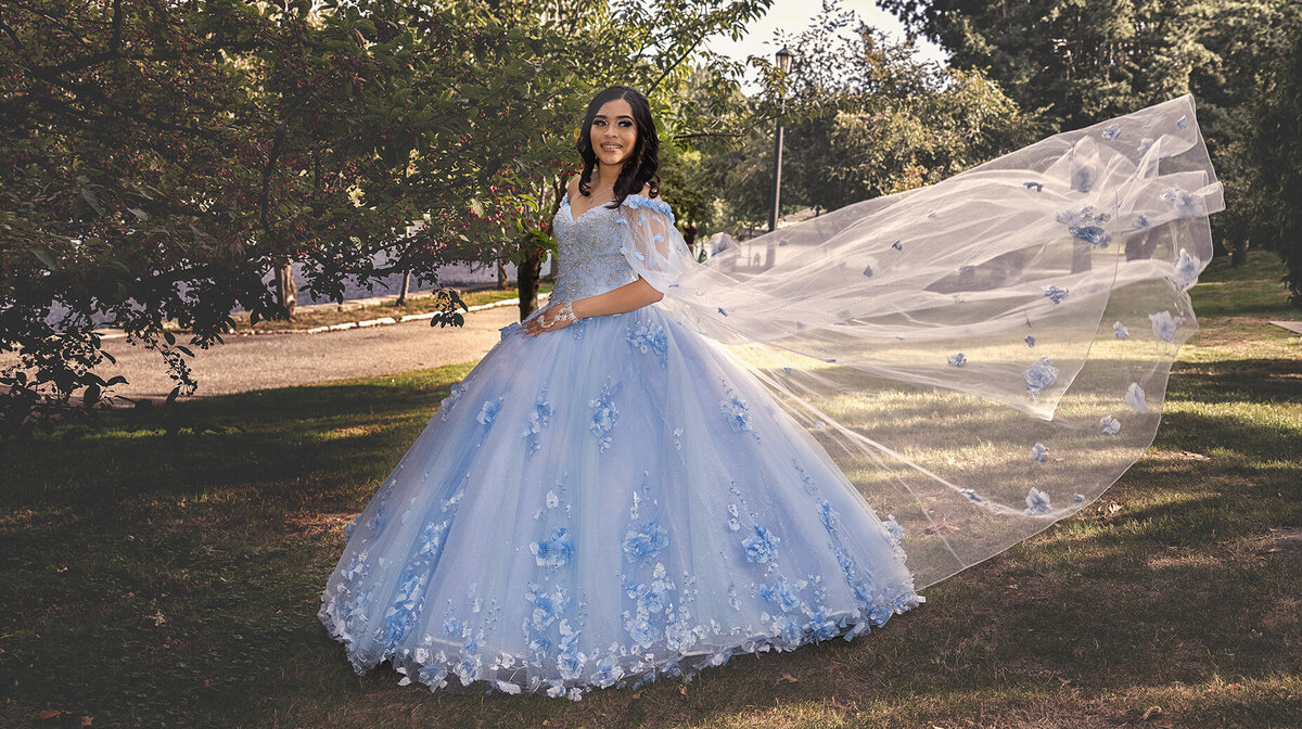 An elegant outdoor portrait of Victoria in her blue quinceañera gown with a flowing cape, captured at University Circle in Cleveland, Ohio. The perfect blend of grace and nature by TheWiz Photography.