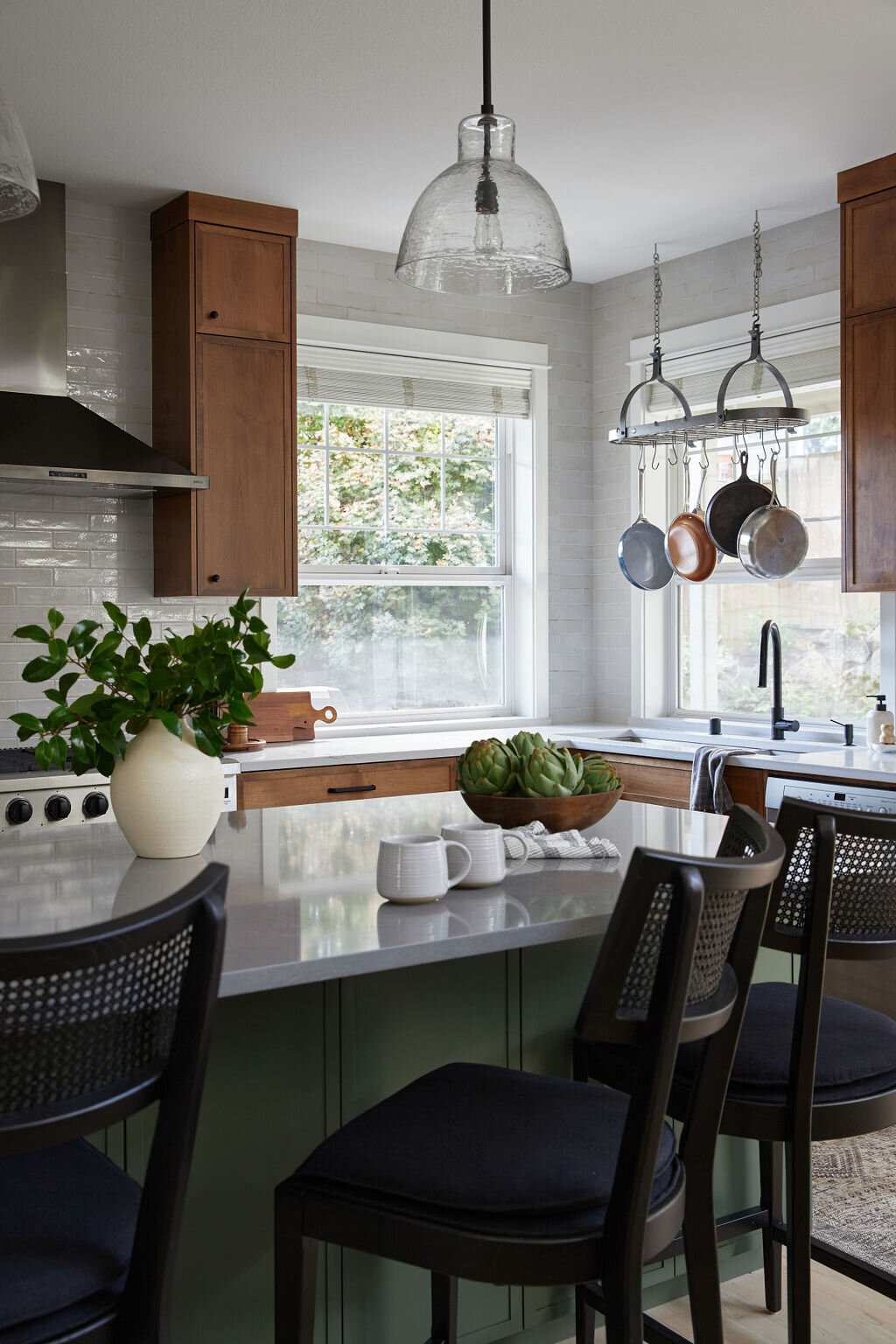 Corner view of a kitchen with two large windows. Perimeter cabinets are natural wood with white countertops, and island is dark forest green with grey countertops. Three black barstools with cane backrests sit in front of the island. A pot rack hangs above the sink. A white vase of greenery, a bowl of artichokes, and two white mugs sit on the island.
