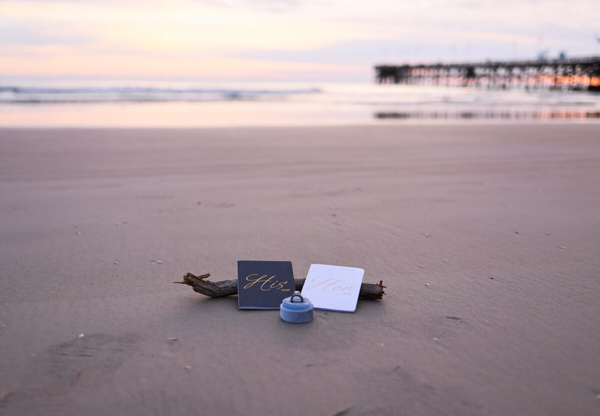 blue groom wedding vow book and white bride vow book with blue ring box with silver wedding ring inside sitting on sand and driftwood