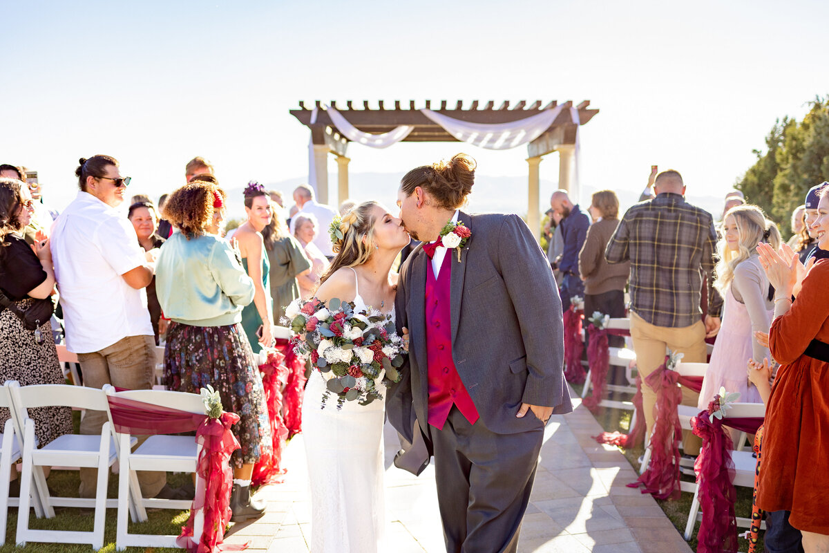 The bride and groom share a kiss as they walk down the aisle after saying “I do,” surrounded by cheering guests and decorated chairs under bright sunlight in Highland, Utah.