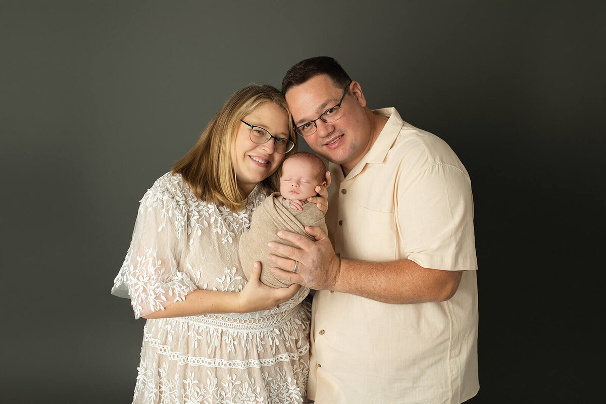 parents proudly holding their swaddled newborn baby boy on a dark grey background in neutral colors for family portraits