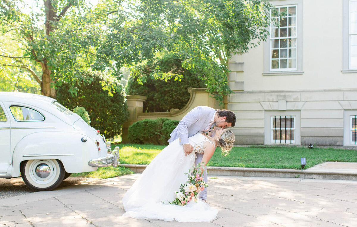 Groom dipping bride in front of Great Marsh Estate and Vintage white car. Taken by Bethany Aubre Photography.